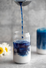 Iced Butterfly Pea Tea Latte on a grey background with flowers. A glass of a cold summer drink. Caffeine free drink. Healthy food concept. Tea is pouring into a glass.