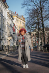 Female model with strong unique style appearance in urban inner city environment walking down street with expensive buildings. Confident black lady stylish red hair with winter good taste fashion