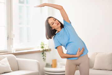 Young pregnant lady doing side bends stretching raised arm indoor