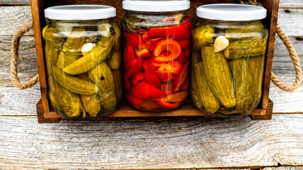 Glass jars with pickled red bell peppers and pickled cucumbers (pickles) isolated in wooden crate. Jars with variety of pickled vegetables. Preserved food concept in a rustic composition. - obrazy, fototapety, plakaty