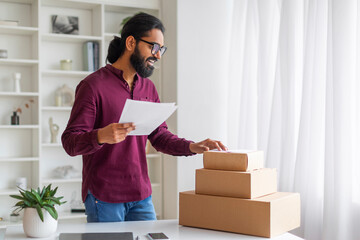 Focused Indian businessman reviewing documents and organizing shipping boxes at home office