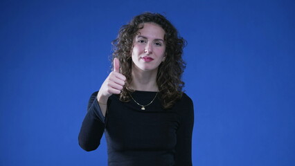 Happy woman showing approval with thumb up, looking at camera while standing on blue background....