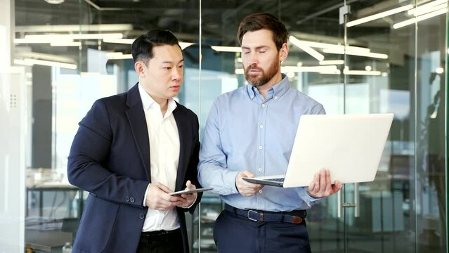 Two diverse colleagues discussing new project, businessmen using digital tablet and laptop compter together in office. professional man corporate employees in conversation working on business project
