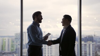 Silhouette of a meeting of two businessmen who shake hands and communicate on a modern background in the office. businesspeople standing against window. Business communication or partnership concept 