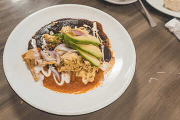 Red enchiladas combined with mole enchiladas, accompanied with chicken, cream, cheese, avocado, on a white plate, on a wooden table.