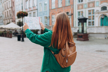 Attractive young female tourist is exploring city. Redhead 30s woman with backpack holding a paper map on city street. Traveling Europe in autumn, rear view