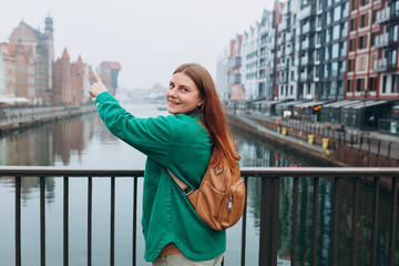 Attractive young female tourist is exploring new city. Redhead woman with backpack pointing finger on city street in Gdansk. Traveling Europe in autumn. Famous Zuraw crane, Motlawa river, rear view