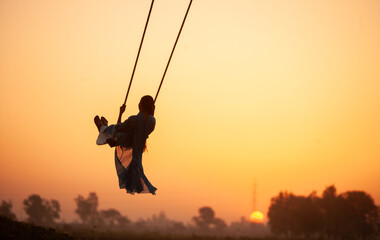 silhouette of a child playing on a swing. A young girl swinging at village during sunset