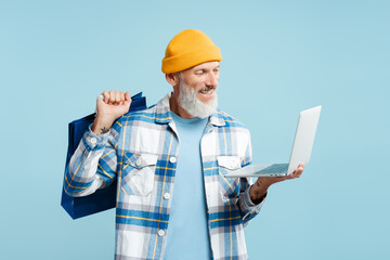 Mature, attractive man wearing stylish shirt and yellow cap holding, using laptop and shopping bag