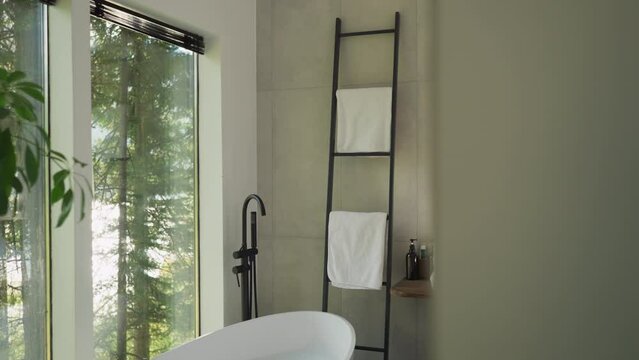 Stylish bathroom and couple brushing teeth. Bathtub and ladder rack by panoramic window in hotel room. Guests of luxury resort do morning routine
