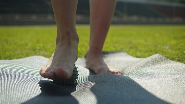 Young woman doing Myofascial Release for feet using a little ball on the sport mat on green grass close up. Massage for legs. Stretching, yoga outdoor