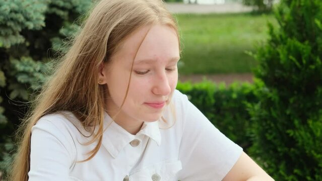 Blonde teenage girl enjoys day in park sitting at table in summer park cafe. Girls radiant smile captured in heartwarming shot