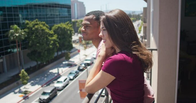 Cute couple smiling and resting on the railing while standing on apartment balcony. Millennial African American man and Caucasian woman on a sunny day. 4k slow motion handheld