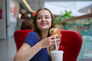 Portrait of young happy woman or beautiful teenager girl eating fast junk food, tasty burger and drink soda in a restaurant or cafe, enjoy meal. 