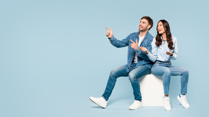 Playful couple in denim pointing outwards, seated on cube, free space