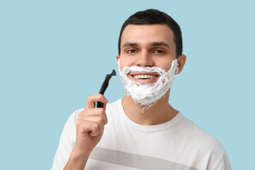 Handsome young man with razor and shaving foam on his face against blue background