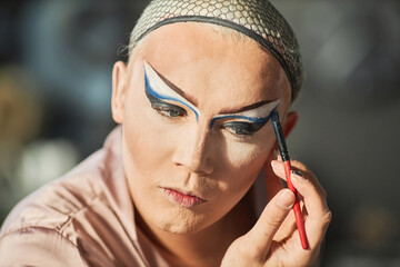 Portrait of drag queen performer doing stage makeup with graphic eyeliner and looking in mirror,...