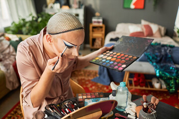 High angle portrait of drag performer doing makeup in messy hotel room and looking in mirror, copy...