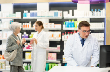 Man pharmacist in pharmacy works with computer, searches among nomenclature of drugs, specifies price and availability of medicine - obrazy, fototapety, plakaty
