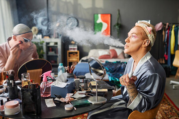 Side view portrait of male drag queen performer blowing smoke clouds while doing makeup in messy hotel room, copy space