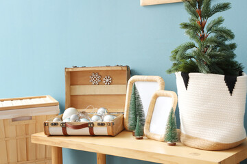 Wooden bench with blank frames, mini Christmas trees and baubles in suitcase near blue wall, closeup