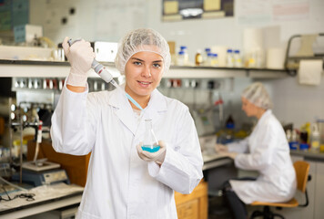 Smiling female chemist pipetting blue reagent into solution in test tube while performing...