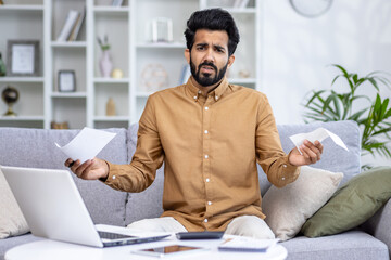 Portrait of upset young Indian man sitting at home on sofa in front of laptop, holding bills and...