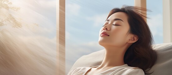 Asian woman resting breathing fresh air feeling mental balance enjoying wellbeing at home on sofa closed eyes put hands behind head relaxing on comfortable sofa in cozy warm light living room
