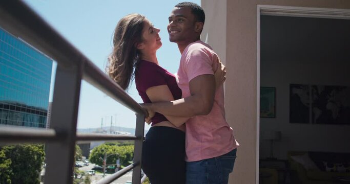 African American and Caucasian couple laughing and holding each other on their balcony in the city. Millennial boyfriend and girlfriend sharing tender moment at home. 4k slow motion handheld