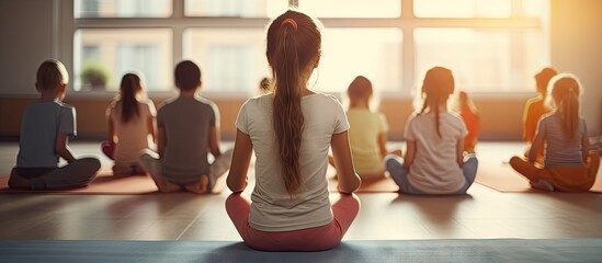 Back view of PE teacher leading Yoga class and meditating with elementary students at school gym. Copy space image. Place for adding text or design