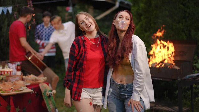 Two girlfriends are standing, looking at the camera, having fun at an outdoor barbecue in the company of friends. A girl blows a bubble gum ball. In the background, a guy plays guitar for his friends.