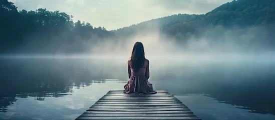 Fotobehang Back view of fashioned young woman sitting on wooden dock looking at view on a misty morning Female hipster with brown hat relaxes on the edge of jetty admiring foggy lake Wonderful nature geta © Ilgun