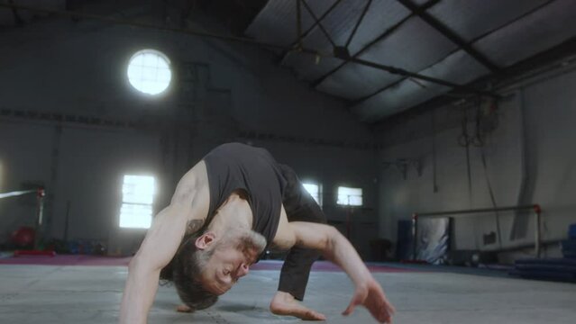 Professional muscular athlete looking at camera and performing acro dance with headstand and floorwork elements during practice in training gym