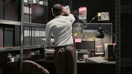 Detective sitting at desk analyzing evidence map on wall, examining law enforcement archive to...