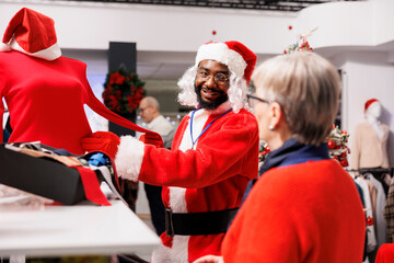 Store employee guiding client in shop, wearing festive santa claus suit while helping customers...