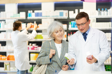 At pharmacy, male chemist consulting mature woman client and shows cream to moisturize of aging skin and convinces doubter to purchase mini version of care product.