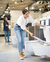 in plumbing department, middle-aged female customer checks strength of toilet seat fasteners before purchasing toilet bowl. buyer pays attention to quality of product