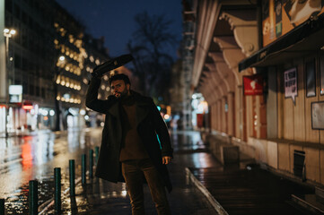 Obraz premium A stylish man waits for a taxi in a busy city at night, covering his head from the rain with waterproof lap top case.