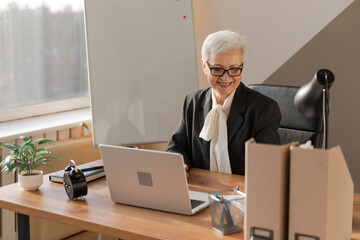 Confident stylish european middle aged senior woman using laptop at workplace. Stylish older mature 60s gray haired lady businesswoman sitting at office table. Boss leader teacher professional worker