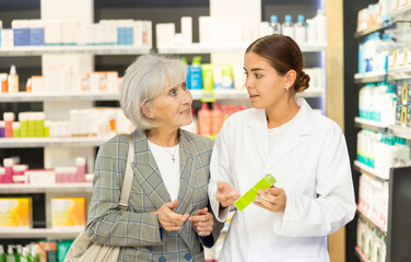 Polite young female pharmacist consulting old costumer about care product in box in chemist's shop