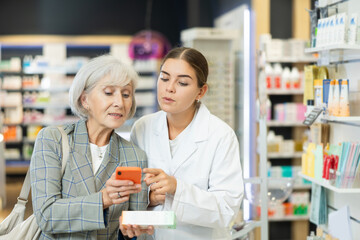 Fototapeta na wymiar Concerned old woman showing to young female pharmacist QR-code of product on her smartphone in chemist's shop