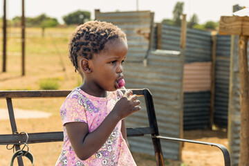 village young african girl eating a lollypop, she is sited on a bench of a donkey cart outdoors