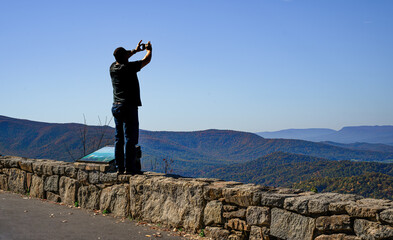 Obraz premium A visitor to Skyline Drive in VA stands on the stone wall to get a better shot of the changing Autumn Leaves and the distant ridge with his cell phone.