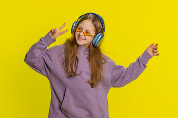 Happy smiling preteen child girl in headphones listening music, dancing disco, fooling around having fun expressive gesticulating hands relaxing on party, making funny moves. Kid on yellow background - obrazy, fototapety, plakaty