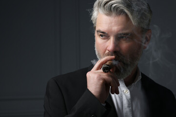 Handsome bearded man smoking cigar against dark grey background. Space for text