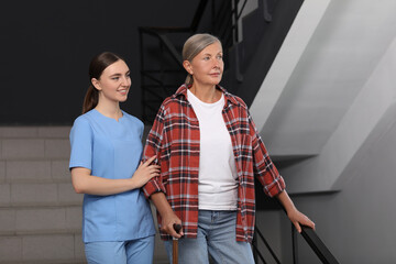 Young healthcare worker assisting senior woman on stairs indoors - obrazy, fototapety, plakaty