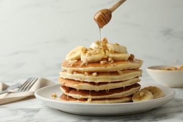 Pouring honey from dipper onto delicious pancakes with bananas and walnuts at white marble table,...