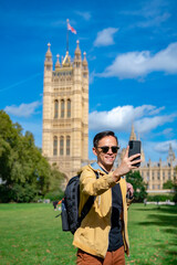 Obraz premium Cheerful young Latin man in casual clothes with backpack in sunglasses and jacket smiling and taking selfie while standing on background of Big Ben against clock tower and cloudy sky in London