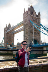 Happy young Latin man wearing sunglasses and red jacket with backpack taking selfie with smartphone near metal railing of famous Tower Bridge in London