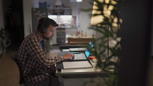 Male American student studies online at home in studio apartment sitting at desk with laptop and taking notes in workbook. Student uses earphones to write notes, watches video online webinar. 